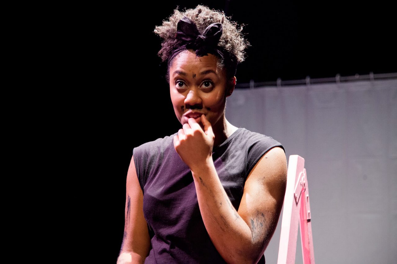 Keisha is a brown-skinned woman looking playfully at an audience out of shot. Her has pen marks all over her body enhancing her natural body hair. She has a moustache. Image Credit: Zuza Photography.