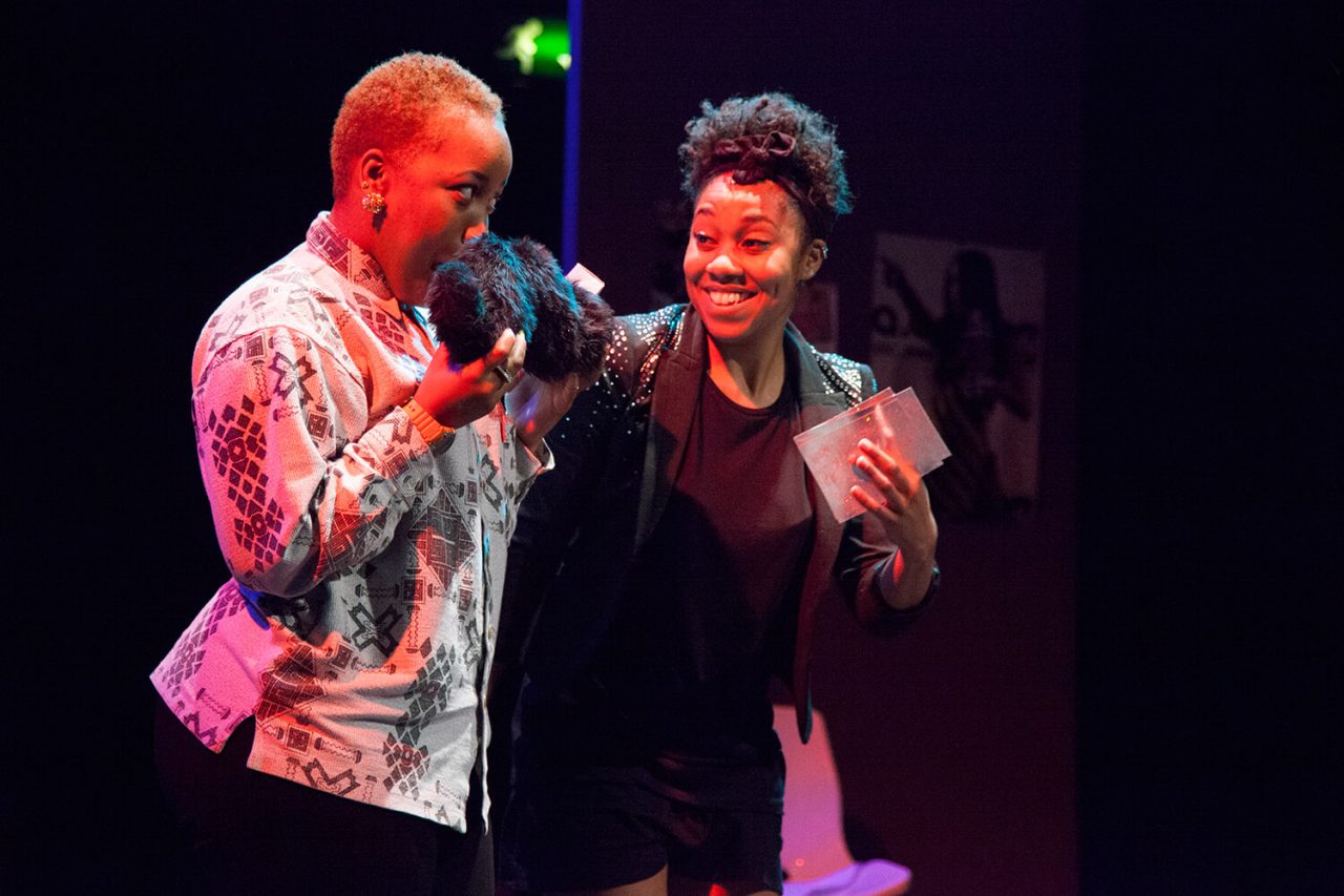 Keisha is a brown-skinned woman wearing a gameshow host blazer. She is stood with a brown-skinned woman holding a comically over-sized moustache to her face. Image Credit - Zuza Photography