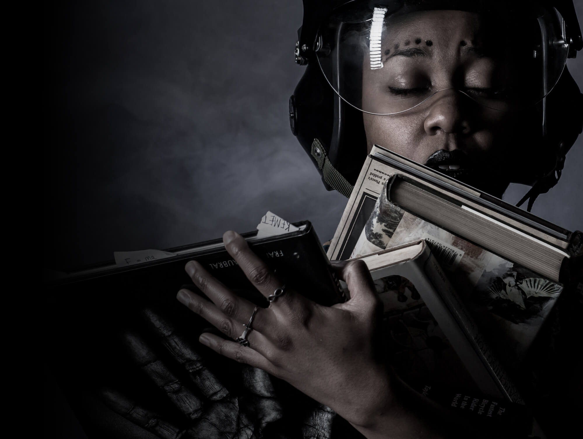 Keisha is wearing a helmet with the moon reflected in it. She has her eyes closed and is clutching several chunky books. Image Credit - Benji Reid