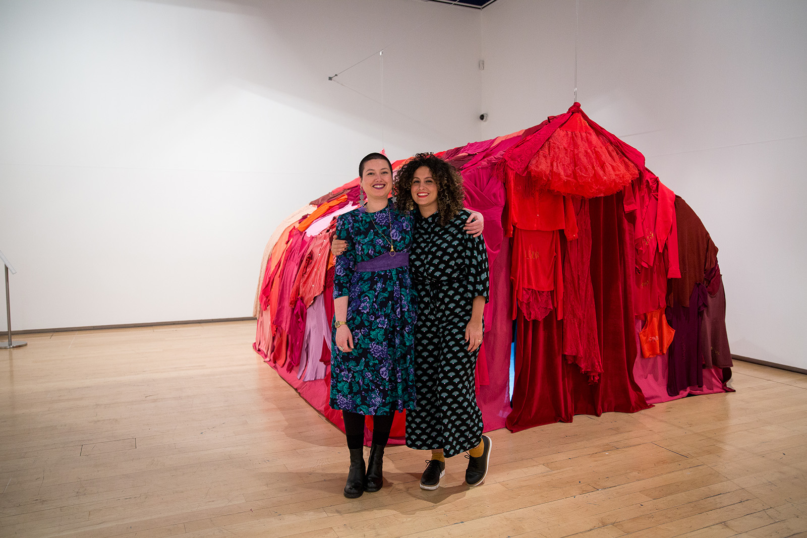 Two brown skinned female artists are stood in front of a tent made of red fabric. Image Credit: Annie Feng