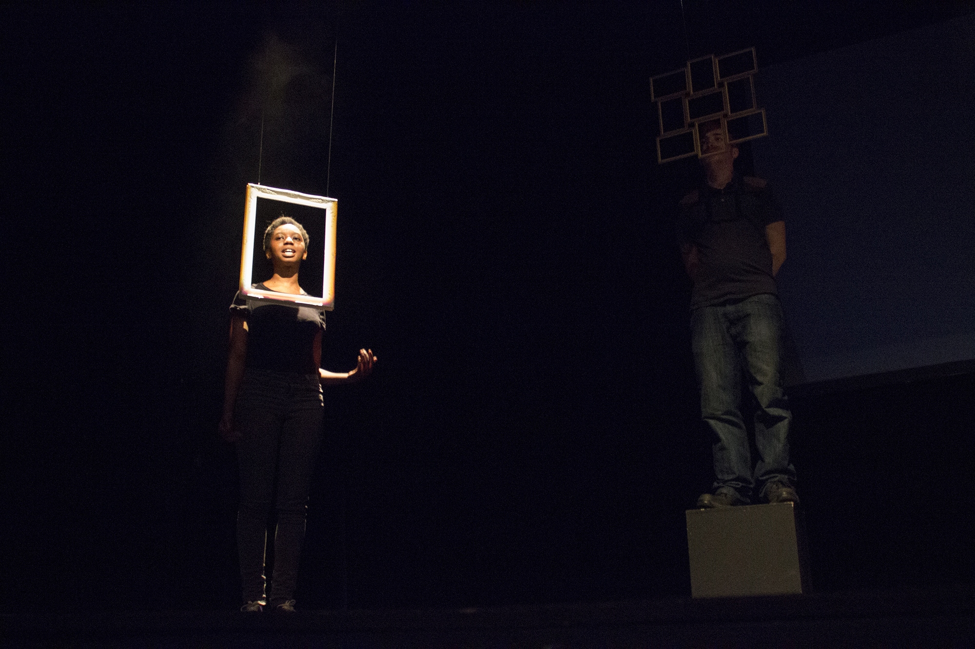 Poet Joan Ncube is stood with her face framed by an empty photo frame. At her left is another poet in the darkness stood in front of more photo frames. Image Credit: Zuzu Photography