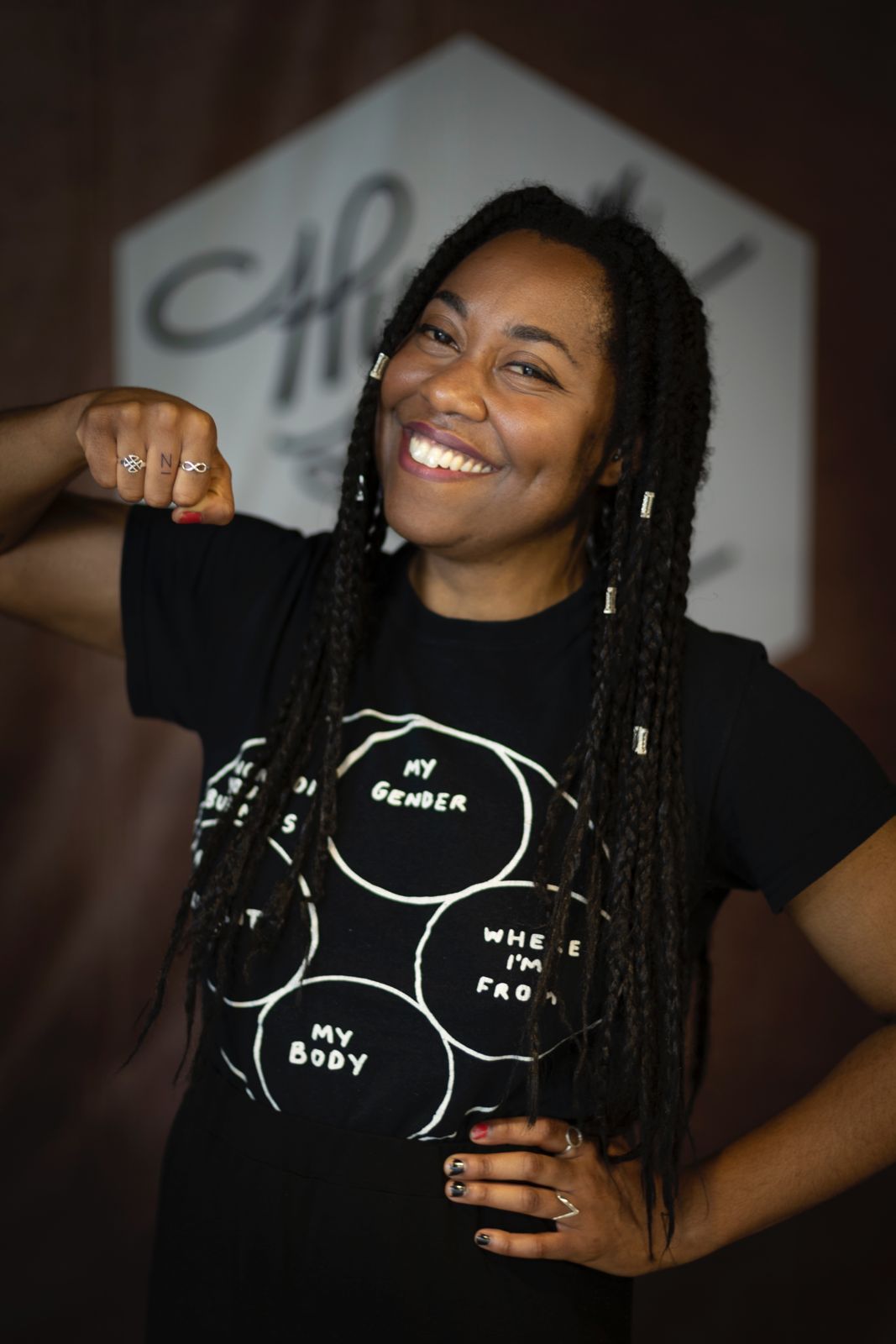 Keisha Thompson is a brown skinned woman smiling with her fist showing a tattoo on her middle finger that is a part of Human Rights Tattoo event. She is wearing a black t-shirt.