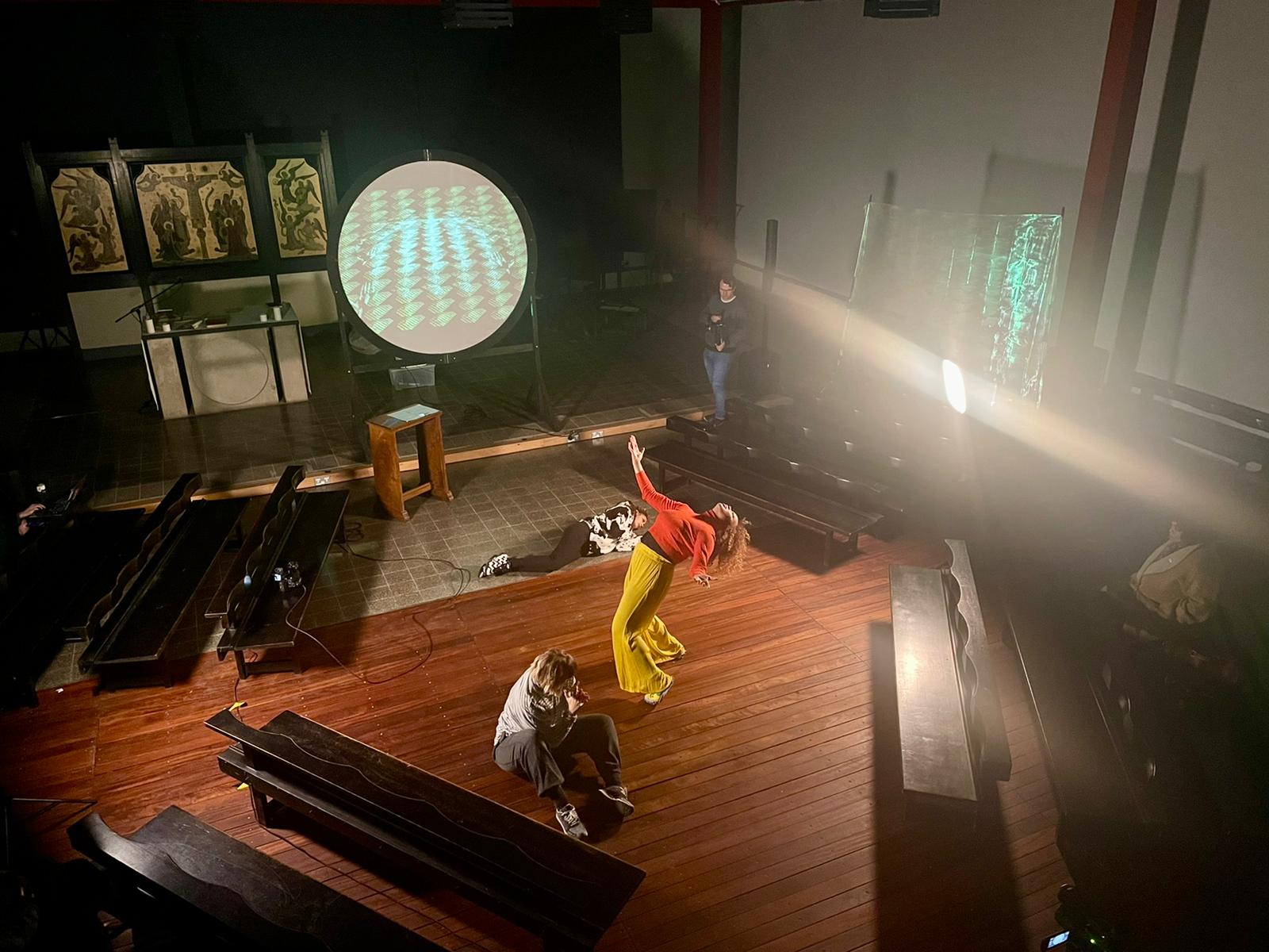 A church hall is lit with white rays and strange blue patterned lighting. A woman wearing orange and yellow is falling backwards with two other woman curled on the floor.