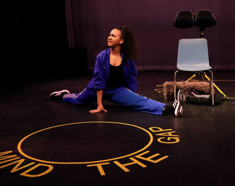 Actress Lauren Fitzpatrick is in the splits position on the floor in an action shot from play, 14%. She is wearing a blue track suit and has long afro hair.