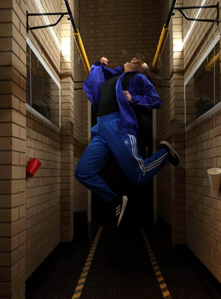 Actress Lauren Fitzpatrick is mid-air in a blue track suit. She is shouting upwards in the confines of small corridor.