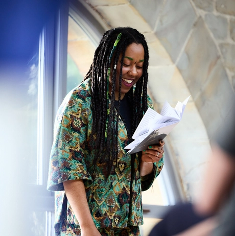 Keisha Thompson is wearing a green suit performing poetry in an archway. She has black braids with green streaks in her hair.