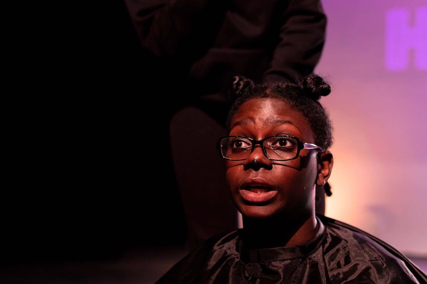 Insert Name, a black woman, sat in a hairdresser seat with a wide-eyed expression